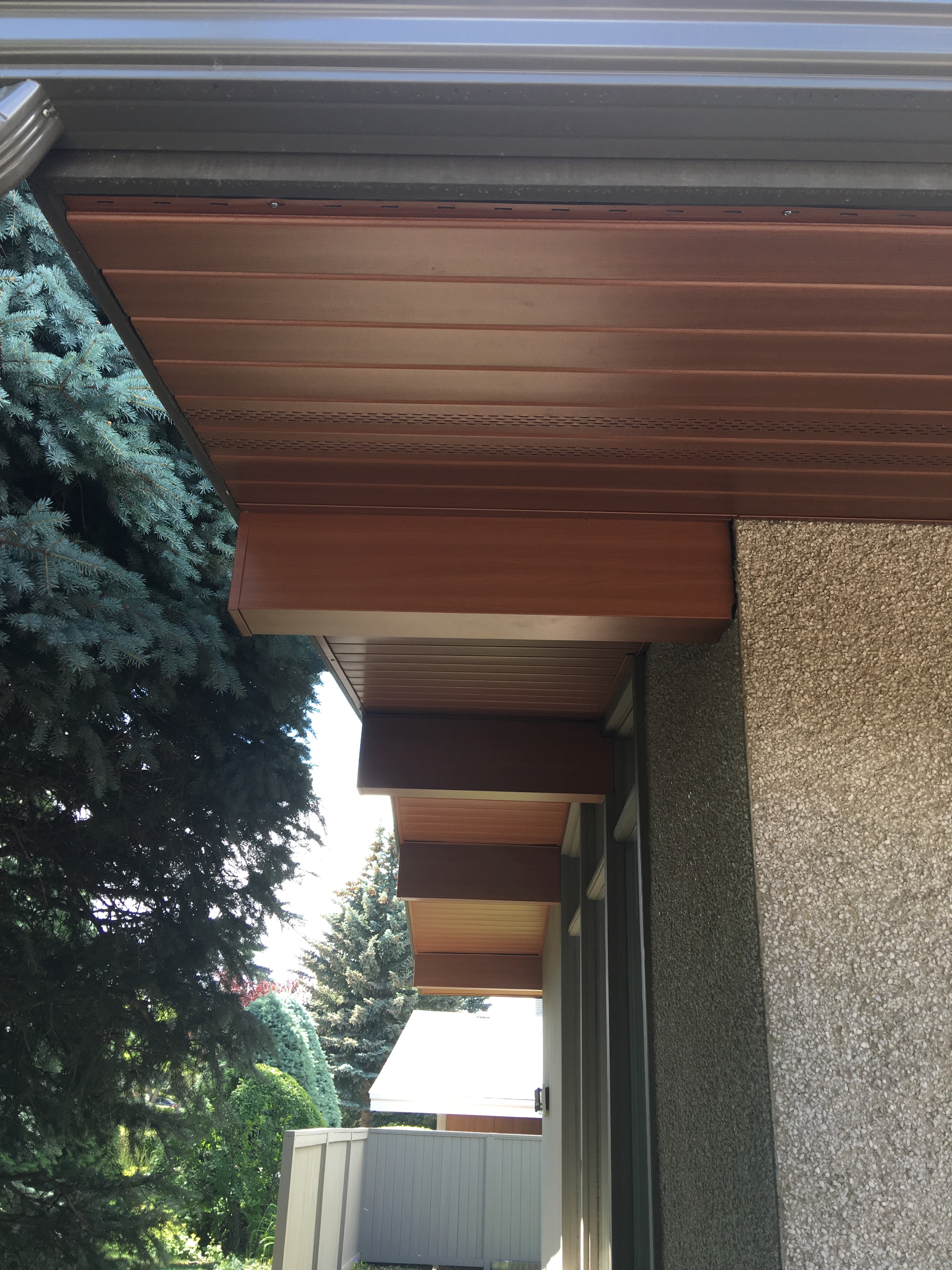 Close-up of modern house soffit with wooden paneling and textured exterior wall, featuring large evergreen trees and a fence in the background.