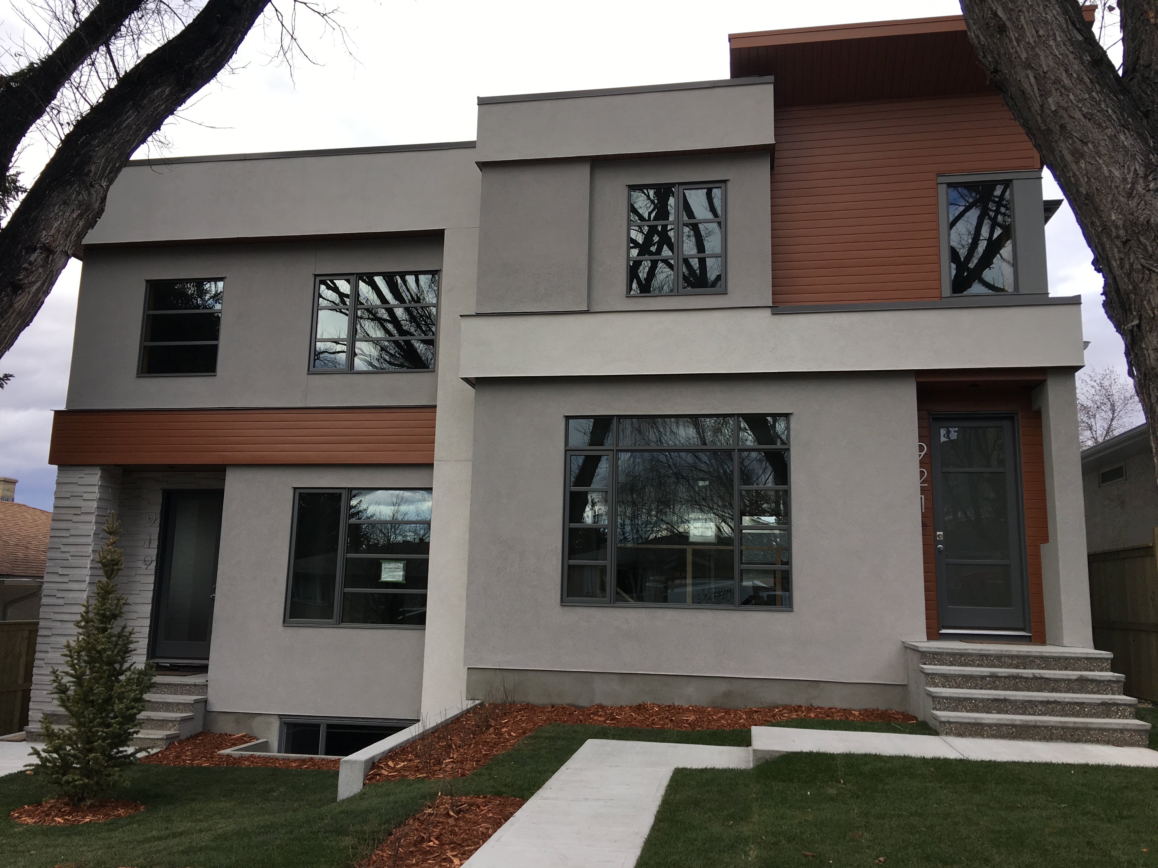 Modern two-story house with large windows, gray and brown exterior, surrounded by trees and newly landscaped lawn.
