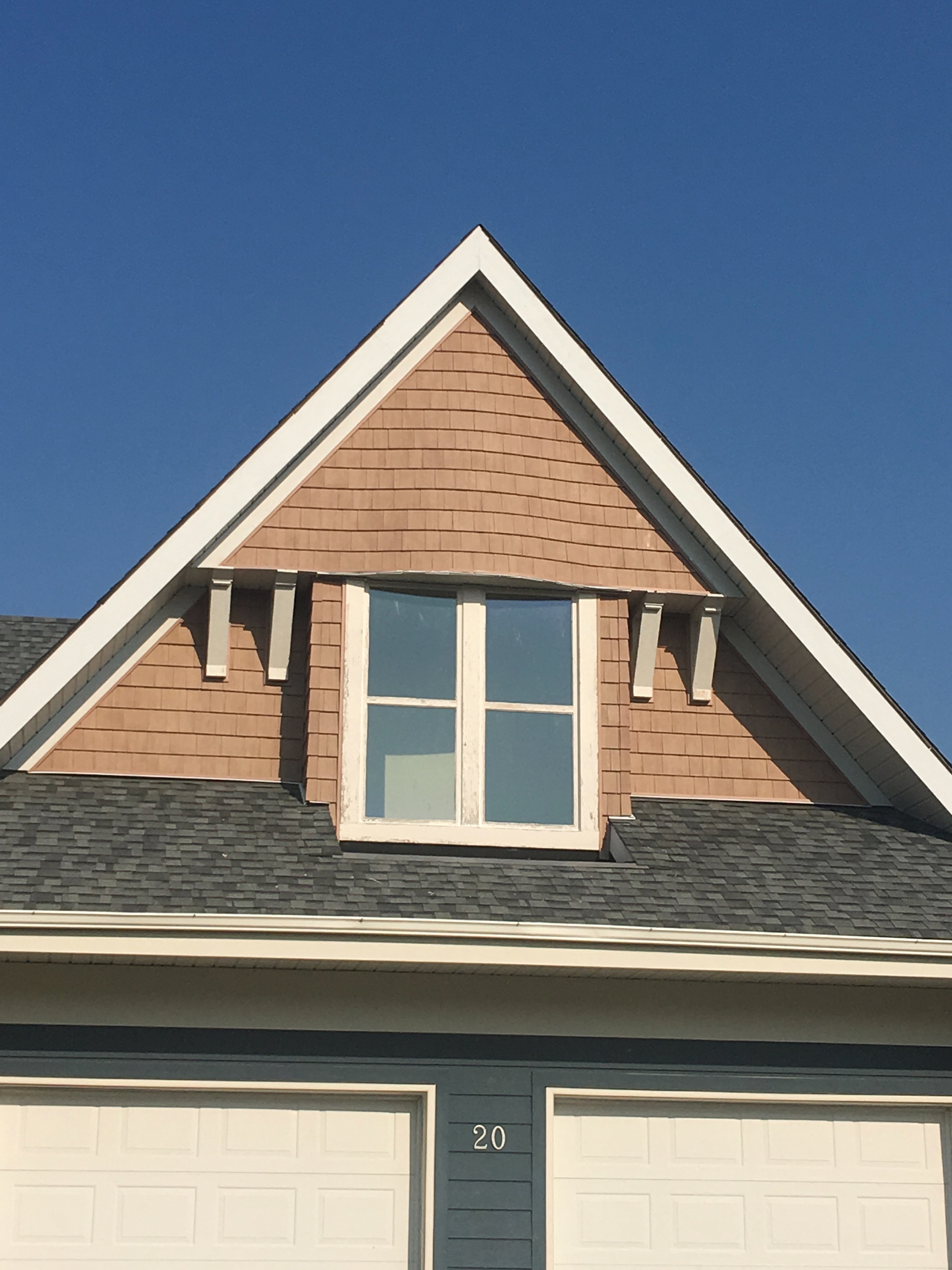 Close-up of a house gable with brown wooden shingles, a double window, and number 20 on the garage below, set against a clear blue sky.